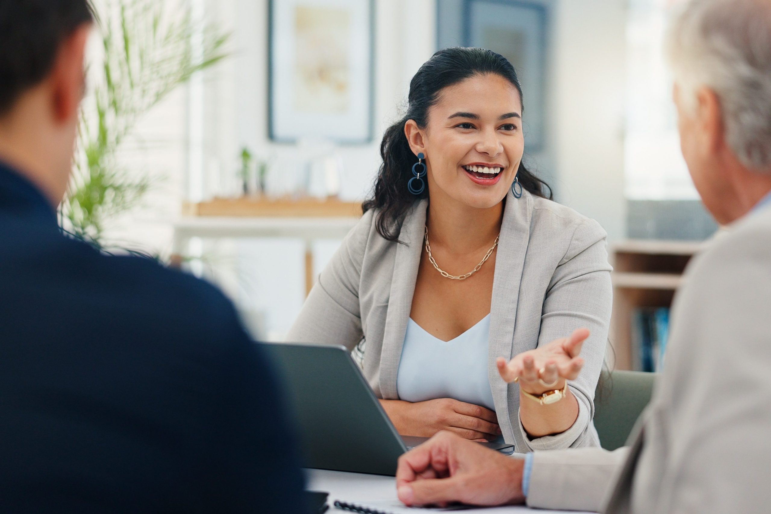 A woman in business attire smiles and gestures while talking to two colleagues at a meeting table with a laptop in front of her.